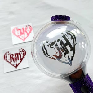 May include: A clear, spherical light stick with a purple jeweled base and top. The light stick features a black and white heart-shaped design with the letter "H" inside. Two heart-shaped stickers are visible in the background.