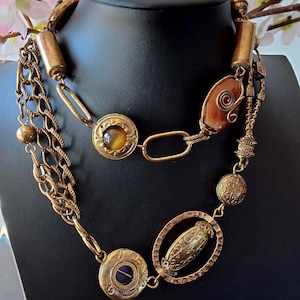 May include: A vintage necklace with multiple strands and decorative elements. The necklace features gold-coloured metal chains, beads, and pendants, including an oval pendant and circular medallions with inset stones. The necklace is displayed on a black mannequin.