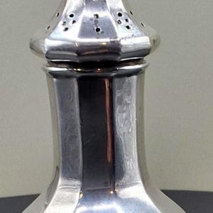 May include: A polished silver-toned salt or pepper shaker featuring a faceted, bulbous body with a narrow neck and a removable domed cap. The cap is designed with a series of small, evenly spaced circular perforations for dispensing contents.