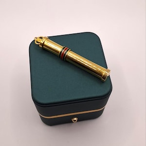 May include: A gold-colored pen with red and black accents rests on a dark green jewelry box. The pen has a crown-shaped top and a cylindrical body. The box is square with gold trim and a small drawer.