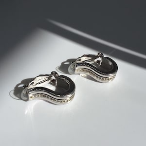 May include: A pair of silver-toned clip-on earrings. Each earring has a curved, layered design with a row of small, clear gemstones. The earrings are on a white surface, with shadows.