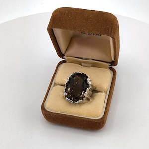 May include: A silver ring featuring a large, oval-cut, dark brown smoky quartz gemstone held by multiple prongs. The ring is displayed inside a brown velvet jewellery box with a cream-coloured interior lining and the text TEL. 277087 printed inside.