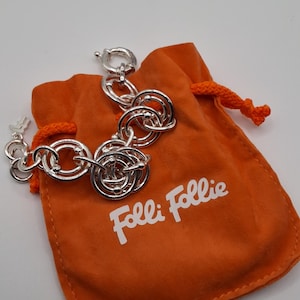 May include: A silver-toned bracelet with a unique, intertwined design rests on an orange drawstring pouch. The pouch features the brand name "Folli Follie" in white lettering. The bracelet's clasp is visible.