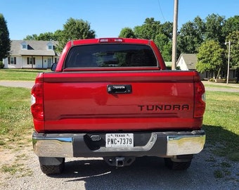 May include: Rear view of a red Toyota Tundra pickup truck. The truck has a chrome bumper, a Texas license plate, and the word "Tundra" in black lettering. The background includes a blue sky, trees, and a house.