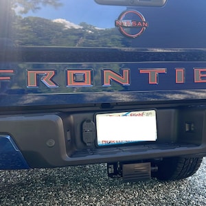 May include: A blue Nissan Frontier PRO-4X pickup truck with a red "FRONTIER" emblem on the tailgate. The truck has a black bumper with a license plate holder and a hitch receiver.