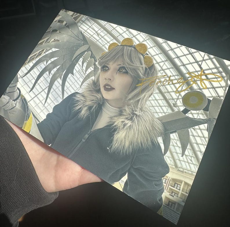 May include: A woman in a grey and black jacket with a fur collar and a gold crown. She has white hair and dark makeup. The background is a blurry image of a building with a glass roof.