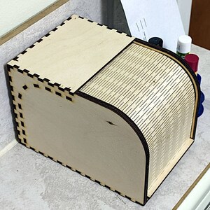 Könnte beinhalten: Eine lasergeschnittene Holzkiste mit Schiebedeckel. Die Kiste besteht aus hellem Holz und weist komplizierte Ausschnitte an den Kanten auf. Die gebogene Seite der Kiste hat ein Muster aus diagonalen Linien. Die Kiste ist teilweise geöffnet.