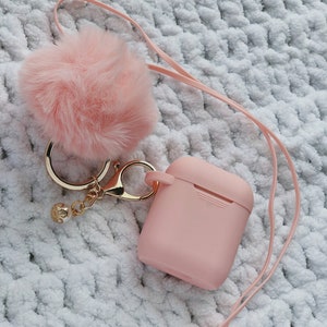 Puede incluir: Un llavero de pompón rosa esponjoso unido a una funda de silicona rosa para auriculares inalámbricos. La funda tiene un anillo de metal dorado y un cordón blanco.