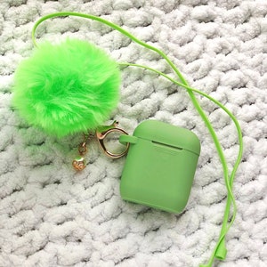 May include: A green fuzzy pom-pom keychain attached to a green silicone case for wireless earbuds. The keychain is connected to the case with a gold metal ring and a small gold charm.