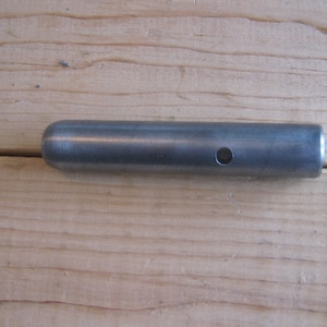 May include: A cylindrical metal object with a rounded end and a small hole. The object is a dull silver color and lies on a light brown wooden surface. The object appears to be a pin or a small rod.
