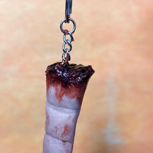 May include: A novelty keychain featuring a severed finger. The finger is pale pink with a dark red, bloody top. It hangs from a silver chain and keyring. The background is a blurred orange and yellow.