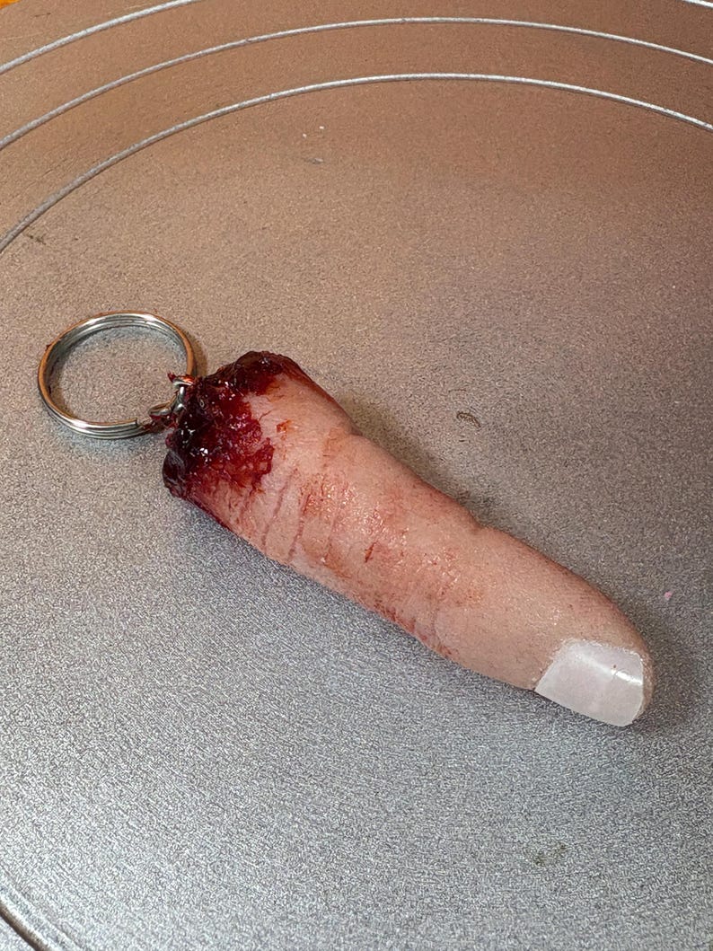 May include: A novelty keychain featuring a realistic, severed finger. The finger is flesh-toned with a white nail and a blood-red, torn end. A silver keyring is attached to the top of the finger.