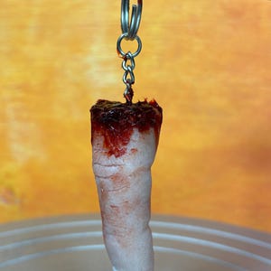May include: A novelty keychain featuring a severed finger. The finger is flesh-toned with a white nail and is covered in fake blood. It hangs from a silver keyring and chain.