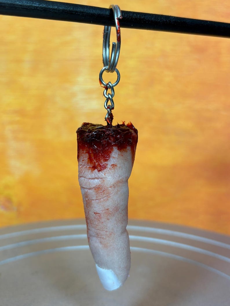 May include: A novelty keychain featuring a severed finger. The finger is flesh-toned with a white nail and is covered in fake blood. It hangs from a silver keyring and chain.