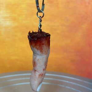 May include: A novelty keychain featuring a severed finger. The finger is flesh-toned with a blood-red top and attached to a silver keyring. The background is a gradient of orange and yellow.