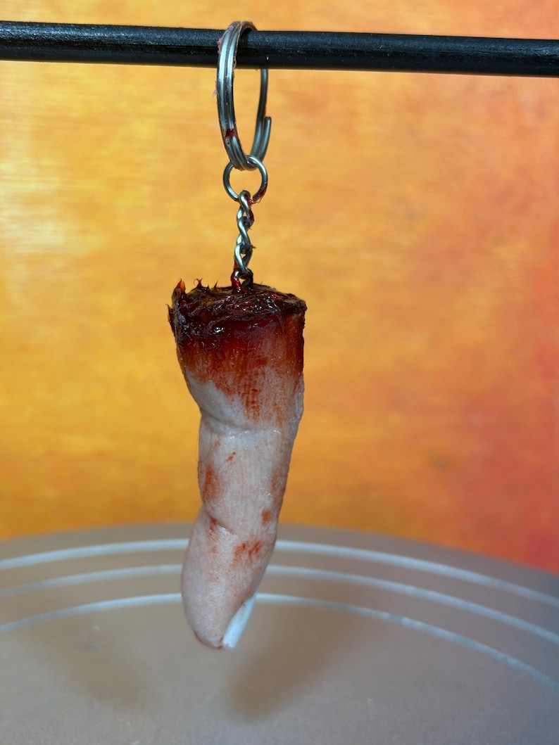 May include: A novelty keychain featuring a severed finger. The finger is flesh-toned with a blood-red top and attached to a silver keyring. The background is a gradient of orange and yellow.