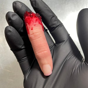 Peut inclure: Un doigt sectionn&eacute; avec de l'os et des tissus expos&eacute;s, recouvert de sang. Le doigt est tenu par une main portant un gant noir. L'ongle est peint avec un vernis transparent.