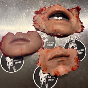 May include: Three prosthetic lips with a realistic appearance, featuring varying shades of skin tones and red accents. Each lip is surrounded by a textured edge, suggesting a torn or damaged effect. The image includes three black and white stickers with the text "DEAD GOAT COMPANY".