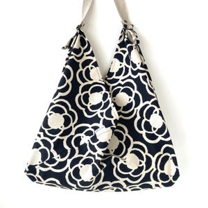 Könnte beinhalten: Eine Schultertasche im Hobo-Stil aus dunkelblauem Stoff mit einem sich wiederholenden, cremefarbenen floralen Linienmuster. Die Tasche hat einen breiten, integrierten Riemen und ein kleines weißes, rechteckiges Etikett mit dem Text "goes on" auf der Vorderseite.