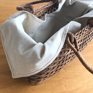 May include: A brown woven wicker basket featuring a natural, light beige fabric liner that drapes over the edges. The basket has two sturdy, braided handles attached to the sides, creating a functional container with a textured, organic appearance.