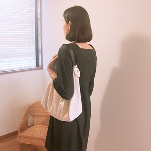 May include: A solid black dress with a square neckline and long sleeves, paired with a large, off-white fabric shoulder bag featuring a dark strap attached with a metal ring. The bag has a soft, gathered texture and hangs at the side.