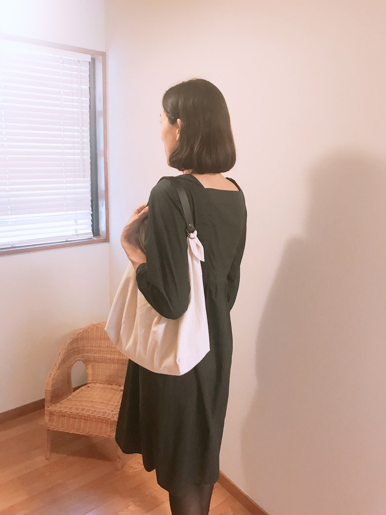 May include: A solid black dress with a square neckline and long sleeves, paired with a large, off-white fabric shoulder bag featuring a dark strap attached with a metal ring. The bag has a soft, gathered texture and hangs at the side.