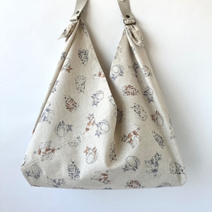 May include: A beige tote bag with a cat print. The bag has a wide, triangular shape and is made of a soft, natural fabric. The cats are drawn in a simple, whimsical style and are scattered across the bag.