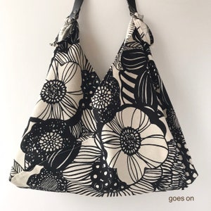 May include: A hobo-style shoulder bag featuring a bold black and white floral pattern. The bag has a triangular silhouette with a black strap attached by metal rings at the top corners. The text "goes on" is printed in the bottom right corner.