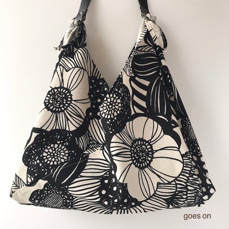 May include: A hobo-style shoulder bag featuring a bold black and white floral pattern. The bag has a triangular silhouette with a black strap attached by metal rings at the top corners. The text "goes on" is printed in the bottom right corner.