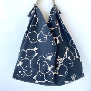 May include: A dark navy blue tote bag with a white illustrated pattern of fruit and birds. The bag has a triangular shape with beige straps and a small pocket. The bag is made of fabric.