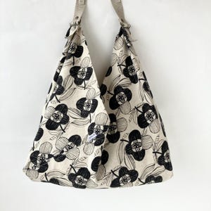 以下が含まれることがあります： 黒い花柄のベージュのトートバッグ。バッグには2本のショルダーストラップがあり、大容量です。花柄のデザインは、様式化された黒い花と葉が特徴です。バッグは丈夫な生地で作られています。