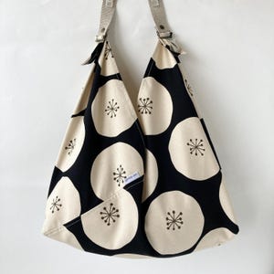 May include: A black and cream-colored tote bag with a repeating floral pattern. The bag features a unique design with two straps and a pocket. The floral pattern consists of large cream-colored circles with a black starburst design in the center.