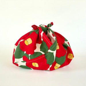 May include: Red and green fabric gift wrap with a floral pattern. The fabric is folded into a pouch shape with a knotted top.