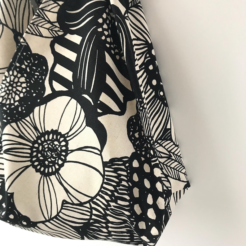 May include: A tote bag made of off-white fabric featuring a bold, high-contrast black graphic print. The pattern consists of large stylized flowers, leaves, and abstract shapes with thick black lines and solid black areas covering the surface.