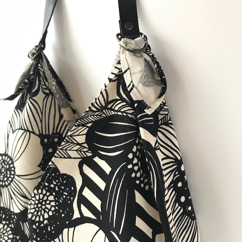 May include: A shoulder bag featuring a bold black and white floral pattern on fabric. The bag is attached to a black leather strap using dark metal rings. The design consists of large, stylized flower motifs and abstract shapes in high contrast.