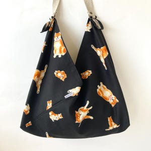 May include: Black tote bag with a repeating pattern of orange and white Shiba Inu dogs. The bag has beige straps and a small pocket. The dogs are in various poses, adding a playful touch to the design.