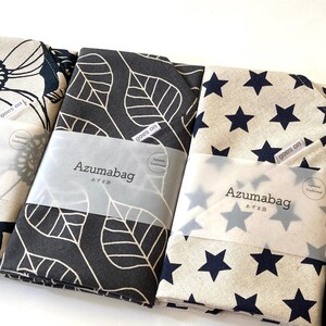 May include: Three folded fabric Azumabag items with distinct patterns: a beige bag with black floral outlines, a black bag with beige leaf outlines, and a beige bag with navy blue stars. Each features a label reading goes on and Azumabag.