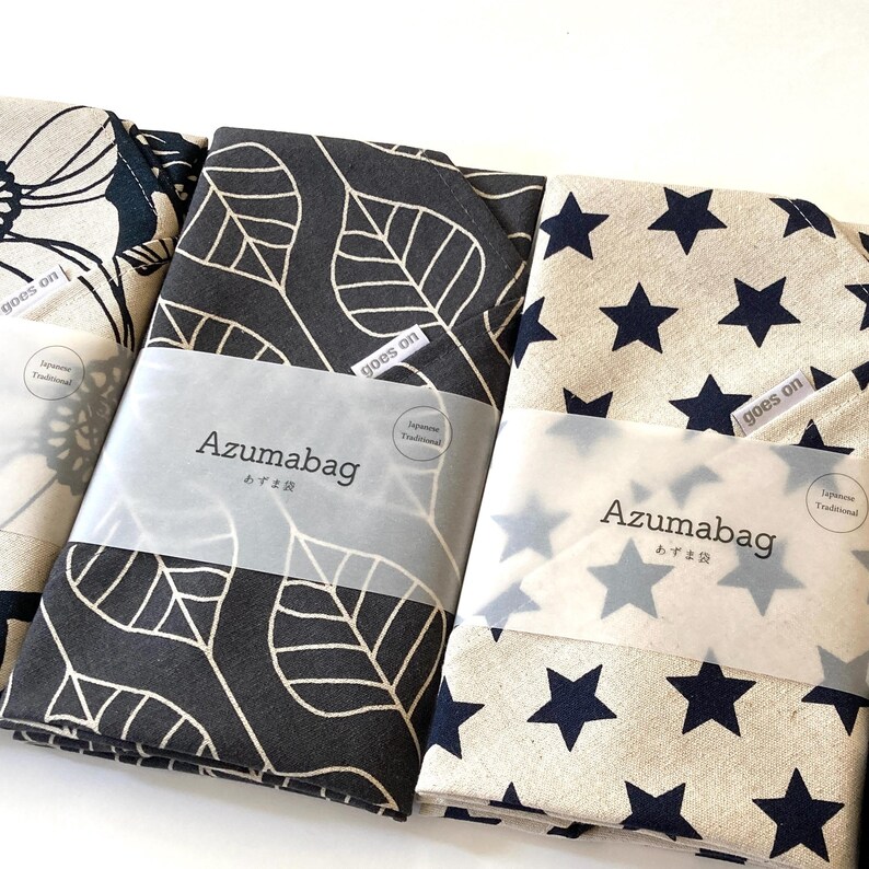 May include: Three folded fabric Azumabag items with distinct patterns: a beige bag with black floral outlines, a black bag with beige leaf outlines, and a beige bag with navy blue stars. Each features a label reading goes on and Azumabag.