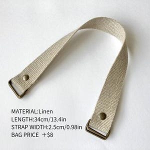 May include: A beige linen bag strap featuring bronze-colored metal hardware at both ends, secured with rivets. The strap measures 13.4 inches in length and 0.98 inches in width. Text on the image specifies: MATERIAL:Linen, LENGTH:34cm/13.4in, STRAP WIDTH:2.5cm/0.98in.