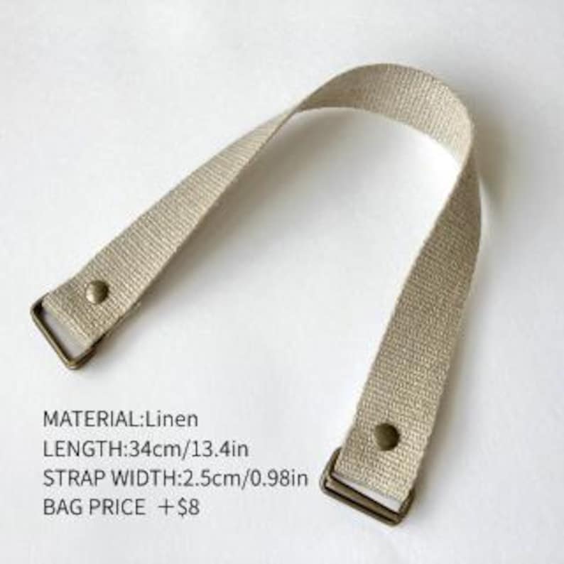 May include: A beige linen bag strap featuring bronze-colored metal hardware at both ends, secured with rivets. The strap measures 13.4 inches in length and 0.98 inches in width. Text on the image specifies: MATERIAL:Linen, LENGTH:34cm/13.4in, STRAP WIDTH:2.5cm/0.98in.