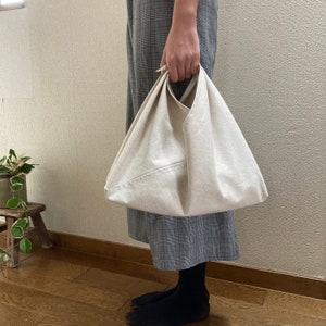 May include: A cream-colored canvas bag with a triangular shape and a single integrated handle. The bag features a folded construction with visible seams and a plain, unpatterned fabric surface, designed to be carried by hand or over the wrist.