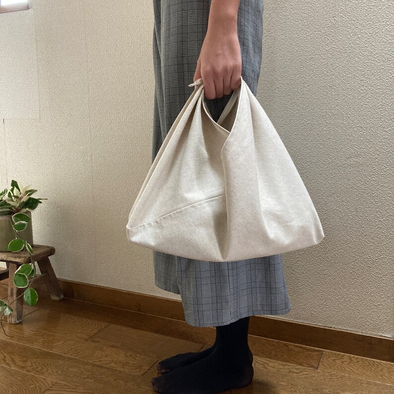 May include: A cream-colored canvas bag with a triangular shape and a single integrated handle. The bag features a folded construction with visible seams and a plain, unpatterned fabric surface, designed to be carried by hand or over the wrist.