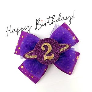 May include: A purple and gold glitter hair bow with a number 2 in the center, shaped like a planet. The bow has purple ribbon with gold stars and a mesh overlay. The words "Happy Birthday!" are written above the bow.