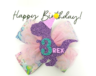 May include: A hair bow featuring a purple glitter dinosaur wearing a pink party hat with a yellow pom-pom. The dinosaur has a teal number 3 and pink text reading REX. The bow is made of pink tulle and patterned fabric. The text Happy Birthday is visible.