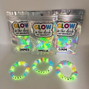 May include: Three glow-in-the-dark DIY bracelet kits in holographic pouches labeled GLOW in the dark DIY BRACELETS with names James, Amelia, and Liam. Three assembled bracelets with colorful beads and black letter beads spelling the same names.