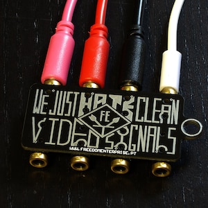 May include: A black metal box with the text "WE JUST HATE CLEAN VIDEO SIGNALS" and the website "WWW.FREEDOMENTERPRISE.PT". The box has four gold-coloured audio jacks and four cables connected to the jacks. The cables are red, pink, black, and white.