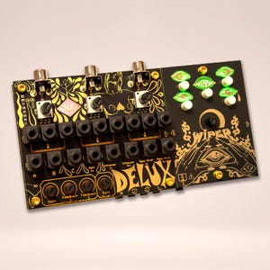 Puede incluir: Sintetizador de música electrónica negro con detalles dorados y la palabra "DELUX" en oro. Cuenta con perillas, conectores y el texto "WIPERS". El diseño incluye patrones y símbolos decorativos.