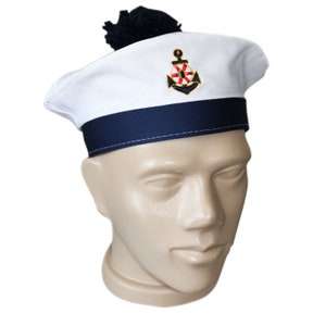 Op de afbeelding: Witte matrozenmuts met een marineblauwe band en een marineblauwe pompon. De hoed heeft een geborduurd ankerembleem met rode en gouden accenten. De hoed wordt getoond op een mannequinhoofd.
