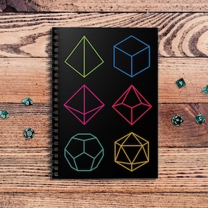 May include: Black spiral-bound notebook with vibrant geometric shapes, such as a pyramid, cube, and dodecahedron. Several green dice are scattered around the notebook on a wooden surface. The notebook is designed for tabletop gaming enthusiasts.