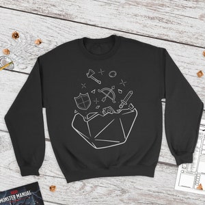 Puede incluir: Sudadera negra con un gráfico blanco de un dado que explota con varios objetos de juego de rol de fantasía, incluyendo una espada, un escudo, un arco y flechas, y un anillo.